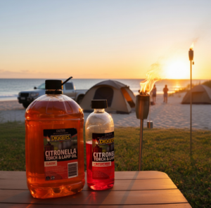 DIGGERS™ Citronella Torch and Lamp Oil bottles on a picnic table at a beach campsite, with a lit tiki torch, tents, and sunset in the background.